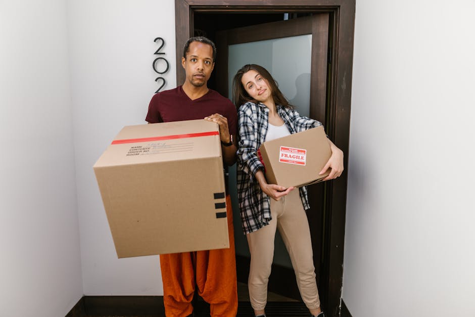 Two individuals standing in a doorway entrance, holding cardboard boxes for home relocation, with packaging labels visible on the boxes. The person on the left, dressed in an orange pair of trousers a