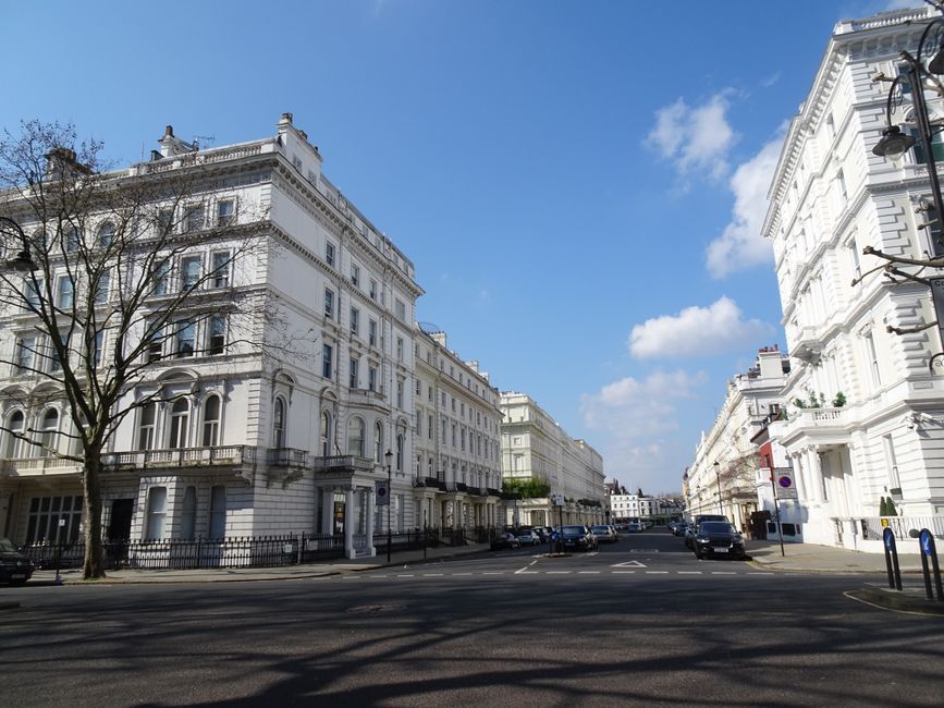 A wide view of a street in an affluent neighbourhood with white, ornate terraced townhouses featuring large windows, decorative cornices, and small balconies. The street has parked cars along both sid